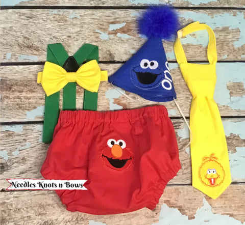 Boys Sesame Street cake smash outfit, each piece made to look like a different character. The diaper cover is red with an Elmo appliqué centered on the back, the bow tie and or tie are yellow with a big bird appliqué and the birthday hat is blue with a Cookie Monster appliqué and the word “one” in white finished off with blue boa at the top of the birthday hat to resemble Cookie Monsters fur. Suspenders are Sesame Street Green.